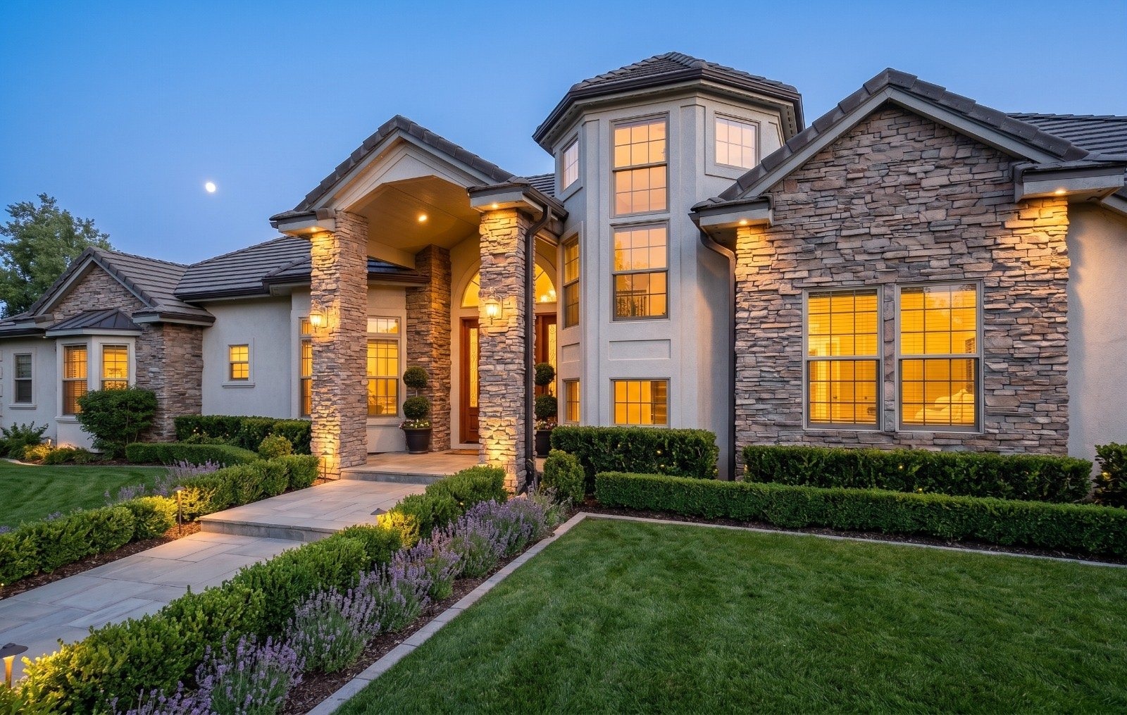 The front of the estate at dusk: stone columns, glowing windows and a lavender-lined path