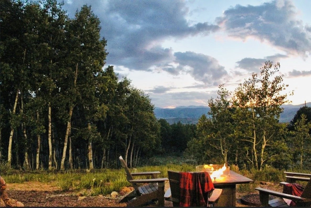 Adirondack chairs around a fire pit at the edge of an aspen forest, with mountains at sunset